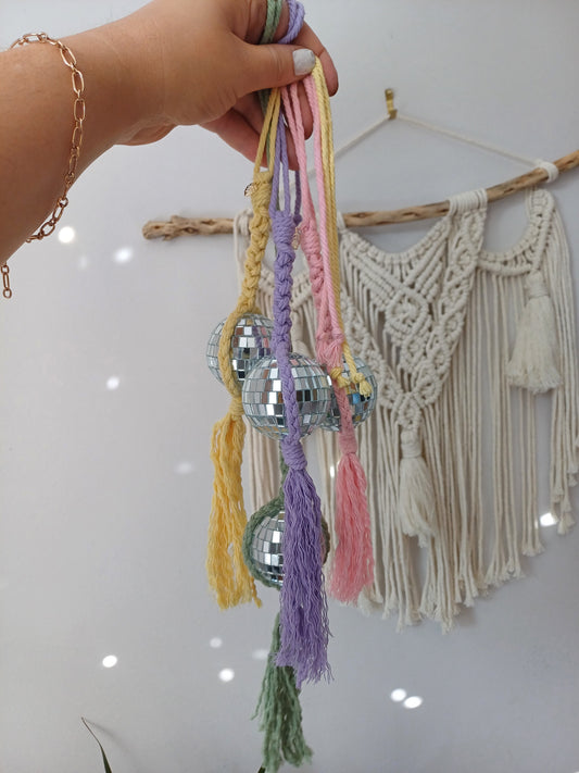 Mini disco ball macrame car charms for with a macrame wall hanging in the background. The disco balls reflects specs of sun light on the macrame wall hanging. There's a pink macrame disco ball and purple macrame disco ball and a green macrame disco ball car accessory. 